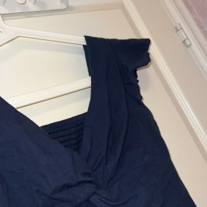 Mörkblå croppad topp med twistad front - Säljer en mörkblå croppad topp med korta ärmar och twistad detalj framtill. Toppen har en smockad rygg och v-ringad halsringning. Perfekt för dig som gillar trendiga och bekväma plagg.Storlek S