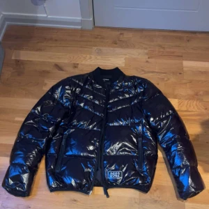 Svart bomber pufferjacka från DSQ2  - Riktigt snygg pufferjacka från DSQUARED2 med den glansiga finishen, lik Moncler Maya men lite mer unikt. Äkta dun, lätt men varm. DSQ2-logga fram. Inga fel eller skador – knappt använd. Storlek: EU 46 – motsvarar ungefär en slim M eller rymlig S. Kvitto finns nypris ca 10000kr