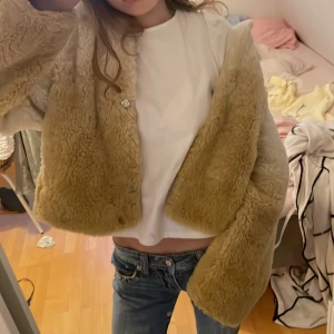 Beige teddyjacka med huva från NA-KD - Säljer en fluffig och mjuk teddyjacka i beige från Weekday. Jackan har huva, långa ärmar och stängs med knappar framtill. Perfekt för att hålla sig varm och snygg under kyliga dagar. Croppad modell som ger en trendig look.hör av dig om du har frågor eller funderingar💕