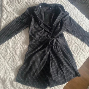 Randig svart jumpsuit från Amisu - Svart playsuit från Amisu med vita tunna ränder, lång ärm och kavajkrage. Snygg omlottkänsla med knyt i midjan för en cool och dressad look. Perfekt för dig som gillar stilrena och trendiga plagg.