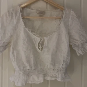 Vit blus med broderade detaljer och volang - Söt vit blus med broderade hålmönster, kort puffärm och volang nertill. Blusen har knytning framtill och resår i midjan för en feminin siluett. Perfekt för vår och sommar!