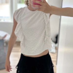 Vit blus med volang från Pieces - Säljer en söt vit blus från Pieces med broderade hål och volangdetaljer längs axlarna. Blusen är ärmlös och perfekt för varma dagar och enkel att matcha med olika nederdelar. Pris går att diskuteras, använd 1-2 gånger!