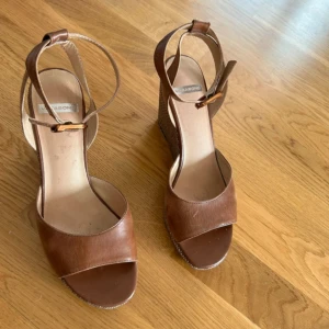 Bruna kilklackade sandaler från Vagabond - Snygga bruna sandaler från Vagabond med kilklack och öppen tå. Skorna har ett brett band över foten och en justerbar rem runt vristen. Perfekta för varmare dagar och ger en stilren look till din outfit.