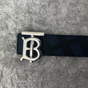 Blått skärp med silverfärgat spänne från Burberry - Säljer ett stilrent blått bälte från Burberry med ett stort silverfärgat spänne i form av Burberrys logotyp. Bältet har diskret mönster och präglad Burberry-text på insidan. Perfekt accessoar för att lyfta din outfit.