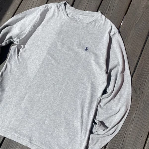 Grå långärmad tröja från Polo Ralph Lauren - Säljer en stilren grå långärmad tröja från Polo Ralph Lauren. Tröjan har en klassisk passform och är tillverkad i mjuk bomull. På bröstet finns den ikoniska broderade loggan i mörkblått. Perfekt för en clean och enkel look. Storlek L (14-16 år)