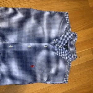 Blårutig skjorta från Ralph Lauren - Klassisk blårutig skjorta från Ralph Lauren med vit- och blått rutmönster. Skjortan har lång ärm, knappar framtill och röd broderad logga på bröstet. Perfekt för en stilren och tidlös look.