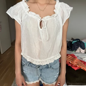 Vit blus med spets och knyt - Supersöt vit blus med korta ärmar, volangkant och spetsdetaljer vid halsringning och nederkant. Blusen har knyt framtill och är lätt och luftig, perfekt för varma dagar. Perfekt skick storlek S.