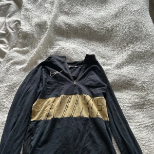 Svart Nike half zip tröja med guldtryck - Svart långärmad tröja från Nike med halv dragkedja och stort guldigt NIKE-tryck över bröstet. Perfekt att träna i.