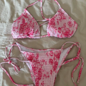Rosa blommig bikini - Supersöt bikini med smockad struktur i ljusrosa och blommigt mönster i starkare rosa. Toppen har trekantsform och knyts i nacken och ryggen, medan trosan har knyt i sidorna. Perfekt för strandhäng och poolparty.