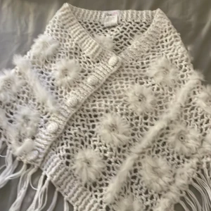 Vit stickad poncho med fransar från Love Forever - Superfin vit stickad poncho från Love Forever med fluffiga blommor och långa fransar. Ponchon har ett öppet mönster och dekorativa detaljer som ger en bohemisk känsla. Perfekt att slänga över en enkel outfit för extra stil.