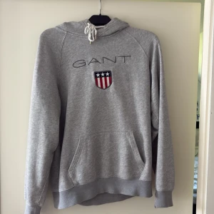 Grå hoddie från GANT - Säljer en grå GANT hoddie, vita snören upptill. Storlek L. Är ett litet hål vid handleden, frågor och fundering bara att kontakta.🤩