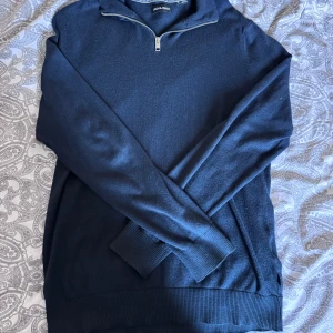 Mörkblå halvzip-tröja från Jack & Jones - Mörkblå långärmad tröja från Jack & Jones med halv dragkedja vid halsen. Tröjan har ribbade muddar vid ärmslut och nederkant samt en klassisk, stilren design. Perfekt för lager-på-lager och enkel att matcha.