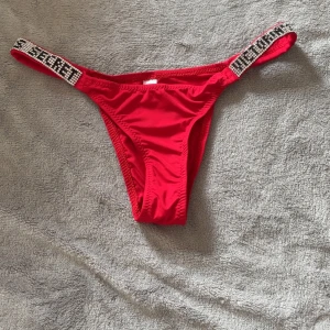 Röd stringtrosa från Victoria's Secret - Säljer en röd stringtrosa från Victoria's Secret med glittriga band där märkesnamnet står i pärlor. Modellen är tanga och har en snygg, enkel design med dekorativa detaljer på sidorna.