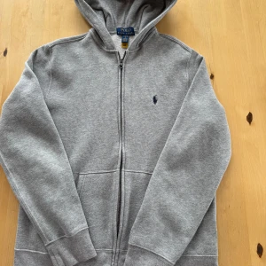 Grå hoodie från Polo Ralph Lauren - Säljer en grå hoodie från Polo Ralph Lauren med dragkedja och huva. Tröjan har broderad logga på bröstet, två fickor fram och är fodrad med mjuk fleece på insidan. Perfekt för kyliga dagar och enkel att matcha med jeans eller joggers.