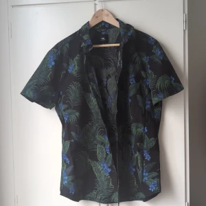 Blommig kortärmad skjorta Clockhouse XL - Snygg svart skjorta från Clockhouse i storlek XL med tropiskt blommönster i grönt och blått. Skjortan har korta ärmar, klassisk krage och knäppning framtill. Perfekt för dig som vill sticka ut med en färgglad och chill stil.