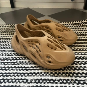 Beige adidas Yeezy Foam Runner - Säljer ett par beige adidas Yeezy Foam Runner med futuristisk design och massor av hål för extra ventilation. Skorna är tillverkade i syntetmaterial och har en chunky, vågig sula. Perfekta för dig som vill sticka ut med en unik och bekväm look.