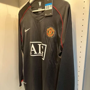 Manchester United långärmad svart fotbollströja från Nike - Säljer en svart långärmad Manchester United fotbollströja från Nike med rött och vitt tryck. Tröjan har klubbmärke på bröstet och AIG som sponsorlogga framtill. Perfekt för fans som vill visa sitt stöd på läktaren eller planen.