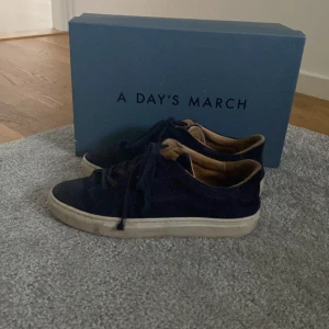 Mörkblå sneakers från A Day's March - Säljer ett par stilrena mörkblå sneakers från A Day's March med skin sulor och klassisk snörning. Skorna har en låg profil och är tillverkade i mocka med innersida i ljusbrunt skinn. Perfekta för en clean och avslappnad look.