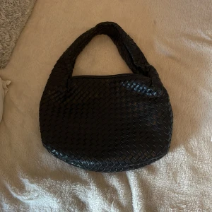 Svart flätad axelväska - Säljer en svart axelväska med flätad design och rundad form. Väskan har ett brett handtag och är perfekt för att bära över axeln. Stilren och enkel att matcha till olika outfits.