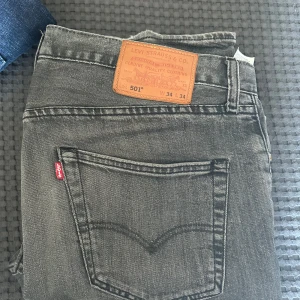 Gråa jeansbyxor från Levi's 501 - Säljer ett par klassiska gråa Levi's 501 jeans med femficksdesign och rakt snitt. Byxorna har den ikoniska läderpatchen bak i midjan och röd Levi's-tag på bakfickan. Perfekta för dig som gillar tidlös denimstil. Hål vid pungen men går att sy ihop!!!!