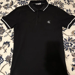 Svart pikétröja Calvin Klein Jeans - Svart pikétröja från Calvin Klein Jeans i slim fit med vita detaljer på krage och ärmslut. Klassisk pikékrage, kort ärm och diskret CK-logga broderad på bröstet. Perfekt för en clean och stilren look.