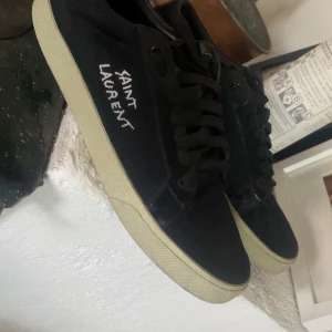Svarta sneakers från Saint Laurent - Svarta sneakers från Saint Laurent med vit sula och handskriven logga på sidan. Klassisk låg modell med snörning och rund tå. Perfekta för en stilren och avslappnad look.