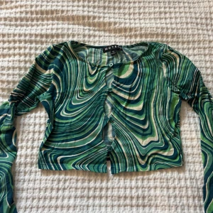 Grön mönstrad topp från Motel - Säljer en  färgstark topp från Motel i gröna och vita vågiga mönster. Blusen har lång ärm, croppad passform och knappar framtill. Perfekt för dig som gillar unika plagg med retrovibbar!