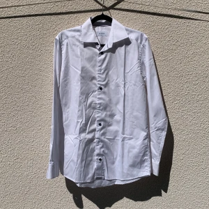 Vit skjorta från Eton - Klassisk vit skjorta från Eton med lång ärm och knappar framtill. Skjortan har en stilren design och är tillverkad i 100% bomull. Slim fit i storlek 39.