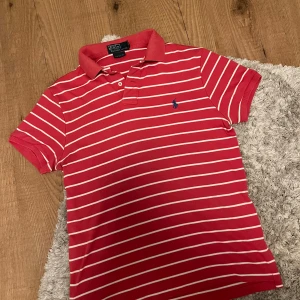 Röd- och vitrandig pikétröja från Polo Ralph Lauren - Säljer en klassisk röd pikétröja från Polo Ralph Lauren med vita ränder och svart broderad logga på bröstet. Tröjan har krage, knappar vid halsen och är kortärmad. Perfekt för en stilren och sportig look. Vill bli av med denna så passar inte pris så kom med bud. 