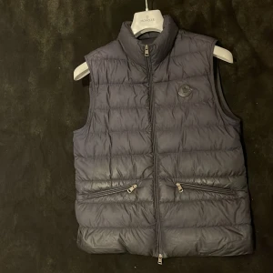 Mörkblå dunväst från Moncler pris kan diskuteras. - Snygg mörkblå dunväst från Moncler med hög krage och två dragkedjefickor framtill. Västen har Moncler-logga på bröstet och stängs med dragkedja. Perfekt för lager-på-lager och stilren look.