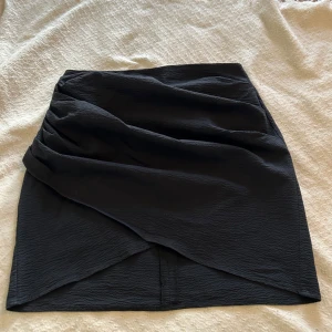 Svart omlottkjol - Säljer en svart kortkjol med omlottdesign och draperad effekt framtill. Kjolen har en stilren look och är perfekt för dig som gillar minimalistisk stil. Materialet känns strukturerat och ger kjolen en snygg form.