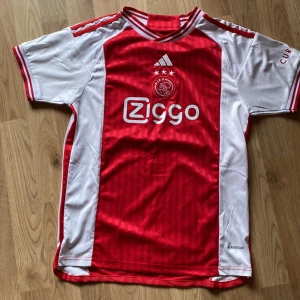 Ajax fotbolls tröja från adidas - Säljer en snygg röd och vit fotbollströja från Adidas med Ajax-logga och Ziggo-sponsortryck. Tröjan är kortärmad och har Aeroready-teknologi för optimal komfort. Perfekt för fotbollsfans!