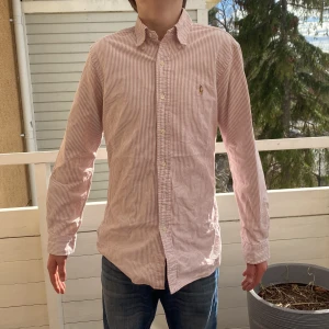 Randig skjorta från Ralph Lauren - Säljer en stilren röd/rosa- och vitrandig skjorta från Ralph Lauren. Skjortan har långa ärmar och en klassisk krage med knappar framtill. Perfekt för en snygg och avslappnad look. Priset är inte fast