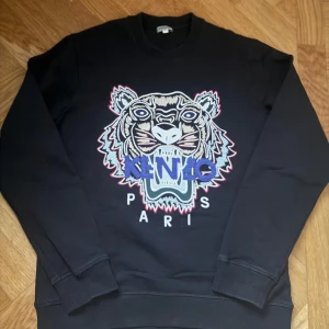 Svart sweatshirt med tigerbroderi från Kenzo - Snygg svart sweatshirt från Kenzo med ett stort, färgglatt tigerbroderi på framsidan. Tröjan har långa ärmar och ribbade muddar vid ärmslut och nederkant. Perfekt för en stilren och trendig look.