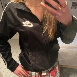 Svart vindjacka från Nike - Säljer en svart vindjacka från Nike med vita detaljer och dragkedja. Jackan har långa ärmar och en vit rand längs ärmarna. Perfekt för en sportig look! Storlek 12-13 år.