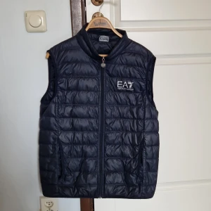 Blå dunväst från Emporio Armani - Snygg blå dunväst från Emporio Armani med dragkedja och broderad logga på bröstet. Perfekt för kyliga dagar och ger en stilren look. Västen har en quiltad design som ger extra värme och komfort.