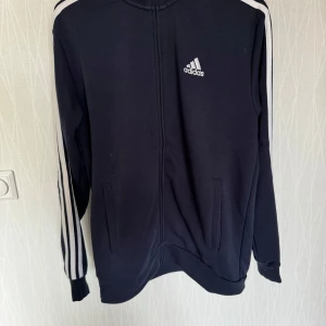 Svart hoodie från Adidas - Säljer en svart hoodie från Adidas med vita ränder längs ärmarna och en dragkedja framtill. Tröjan har långa ärmar och en hög krage. Perfekt för träning eller vardagsbruk.