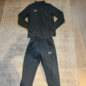 Under Armour dress - Snygg grå träningsdräkt från Under Armour med dragkedja på jackan och elastisk midja på byxorna. Perfekt för träning eller en avslappnad dag. Diskret logotyp på både jacka och byxor. Storlek S i byxor och tröja, personen på bilden är 180cm. Dressen köpt för 1000kr!