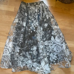 Blommig långkjol i svart och vit - Säljer en elegant långkjol med ett vackert blommigt mönster i svart/  grå/ vit och dragkedja bakom . Kjolen har en transparent överlager och ett svart varmt lager under funkar bra för vår eller höst. 