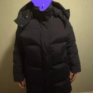 Woolrich Svart Dunjacka - Säljer en svart dunjacka från Woolrich med huva. Jackan är lång och har en klassisk design med dragkedja framtill. Perfekt för kalla vinterdagar. Inte använd.