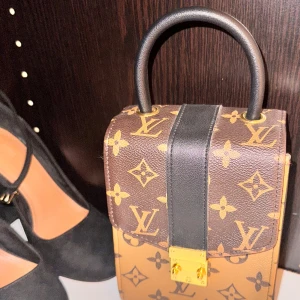 Brun och svart handväska  - Snygg handväska i brunt och svart med  gulddetaljer. Väskan har ett handtag och en dekorativ låsdetalj framtill. Perfekt för att ge en lyxig touch till din outfit.