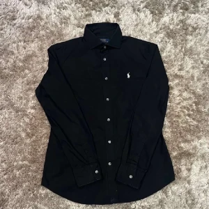Svart skjorta från Ralph Lauren - Snygg svart skjorta från Ralph Lauren med klassisk krage och vita knappar. Skjortan har en broderad logotyp på bröstet och långa ärmar. Perfekt för en stilren look. Pris kan diskuteras 