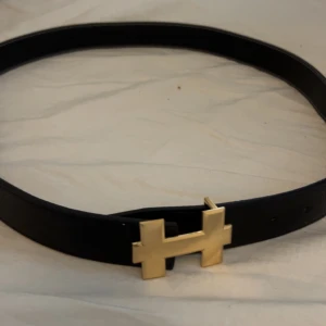 Svart bälte med guldfärgat spänne - Elegant svart bälte med ett stilrent guldfärgat spänne i form av ett H. Perfekt för att ge en lyxig touch till din outfit.