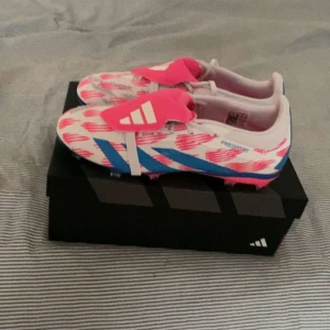 Adidas fotbollsskor med rosa och blå detaljer - Snygga Adidas fotbollsskor i vitt med rosa och blå detaljer. Skorna har en modern design med snörning och är perfekta för fotbollsplanen. De har en rund tå och är gjorda för att ge bra grepp och komfort under spelet.