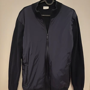 Mörkblå cardigan från John Henric - Snygg mörkblå bomberjacka från John Henric med dragkedja framtill. Jackan har långa ärmar och en stilren design, perfekt för en modern look. Den är tillverkad i ett syntetmaterial som ger en skön passform. Strorlek m men sitter som Small