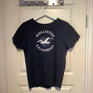 Mörkblå t-shirt från Hollister - Säljer en mörkblå t-shirt från Hollister med tryck av en fågel och texten 'Hollister California' på framsidan. Perfekt för en avslappnad stil. T-shirten har korta ärmar och rund hals. Jätte skön och härlig på sommaren, och fin till vita linnebyxor.