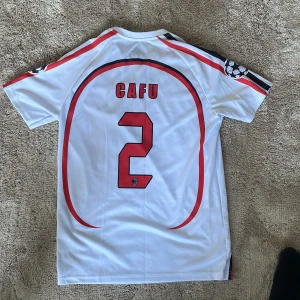 Vit AC Milan fotbollströja med Cafu 2 - Säljer en vit AC Milan fotbollströja från Adidas med Cafu och nummer 2 på ryggen. Tröjan har röda och svarta detaljer samt korta ärmar. Perfekt för fotbollsfans!