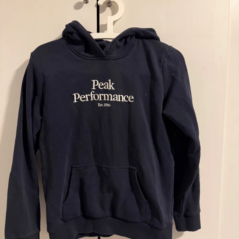 Säljer en snygg mörkblå hoodie från Peak Performance med vit logga på bröstet. Tröjan har en klassisk känguruficka och en bekväm huva. Perfekt för en avslappnad stil.. Neuletakit & Villapaidat.