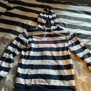 Race marine - Säljer en randig hoodie i marinblått och vitt med tryck på bröstet. Tröjan har en stor ficka framtill och justerbar huva med snören. Perfekt för en avslappnad stil.