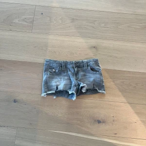 Grå jeansshorts med fransar  - Säljer ett par grå jeansshorts med fransiga kanter och snygga bakfickor med knappar. Perfekta för en avslappnad sommarlook. De har en klassisk femficksdesign och knappgylf.